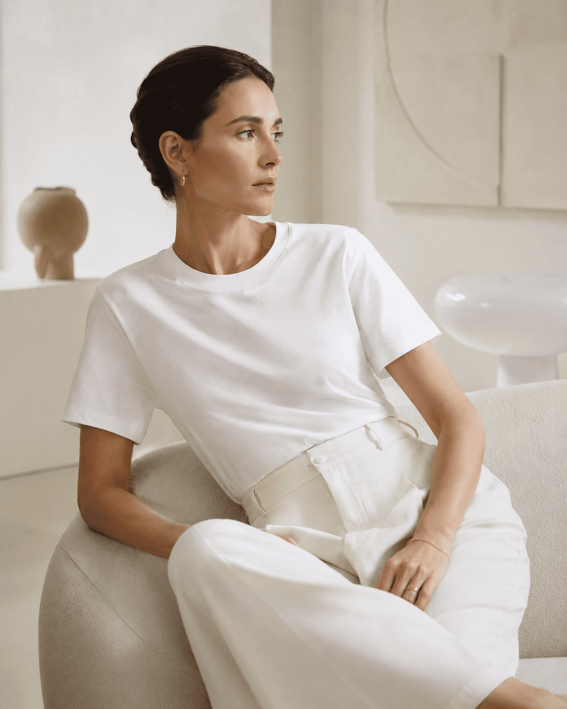 Woman seated in a white T-shirt and cream wide-leg trousers in a softly lit minimal interior